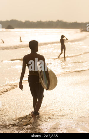 Man Walking am tropischen Strand von Midigama, Sri Lanka an sunsen 4 mit Surfbrett in der Hand. Stockfoto