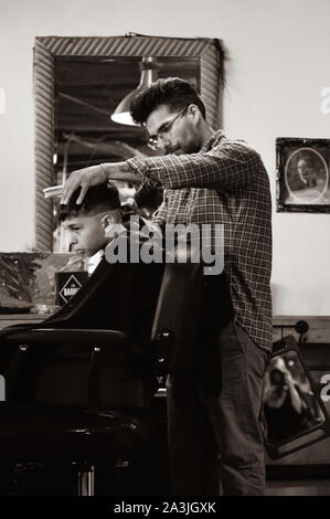 Ein Junge mit Schürze sitzt in Barbier Stuhl, während mit hip Haarschnitt in trendigen '81 Friseure shop in Tucson, AZ, in sepia Ton Stockfoto