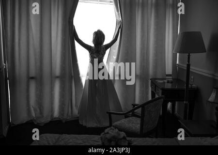 Die blonde Braut in einem Boudoir Kleid öffnet die Vorhänge im Zimmer und schaut aus dem Fenster während der Vorbereitung für die Hochzeit und warten auf die Stockfoto