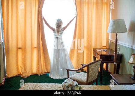 Die blonde Braut in einem Boudoir Kleid öffnet die Vorhänge im Zimmer und schaut aus dem Fenster während der Vorbereitung für die Hochzeit und warten auf die Stockfoto