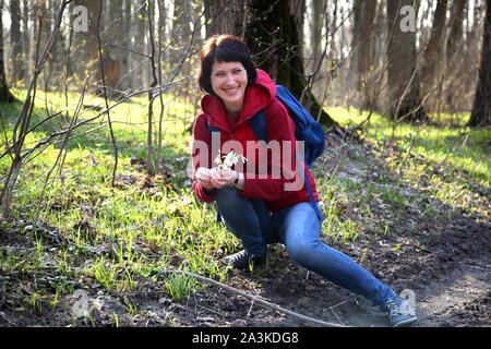 Ein Mädchen sammelt Schneeglöckchen im Wald. Schöne junge Frau taucht in den Strahlen der Frühlingssonne auf einer Lichtung im Wald. Stockfoto