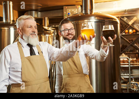 Zwei Spezialisten stehen mit Gläsern Bier, Gläser suchen und lächelnd. Brauerei Arbeitnehmer in weißen Hemden und braun Schürzen Brauerei Hintergrund posiert. Konzept der Handwerk Bier Brauerei. Stockfoto