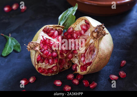 Frische natürliche Granatapfel fuit und einige rote Körner auf einem schwarzen Holztisch neben einigen grünen Blätter in eine rustikale Küche. Leere Raum für Editor Stockfoto