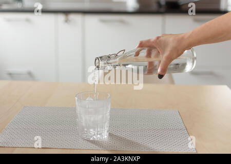 Weibliche Hand gießen kaltes Wasser aus Flasche in Glas mit Eiswürfeln auf hölzernen Tisch über Küche Hintergrund. Stockfoto