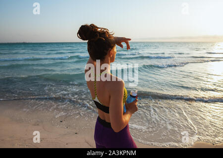 Hinter gesunden Sport Frau in Fitness Kleidung mit einer Flasche Wasser in die Ferne schauen, der am Ufer des Meeres am Abend gesehen. Stockfoto