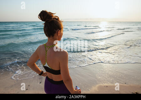 Hinter der jungen Frau im Sport Sport Style Kleidung mit einer Flasche Wasser in der Ferne auf das Meer Küste bei Sonnenuntergang gesehen. Stockfoto