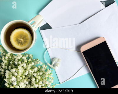 Zusammensetzung der Tasse Tee, Smartphone, leeren Umschlag Karte und Maiglöckchen Blumenstrauß auf Th pastell-blaue Hintergrund. Ansicht von Oben. Stockfoto