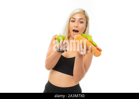 Ein junger Sport Mädchen mit blondem Haar in einem schwarzen Sport top und schwarze Leggings hält einen grünen Apfel und Sandwich. Stockfoto