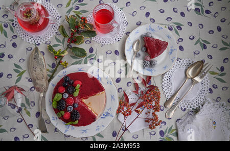 Käsekuchen mit Himbeeren und Brombeeren, Heidelbeeren, Himbeeren und Minze garniert. Auf einem bunten Tischdecke, Blätter und Beeren. Stockfoto