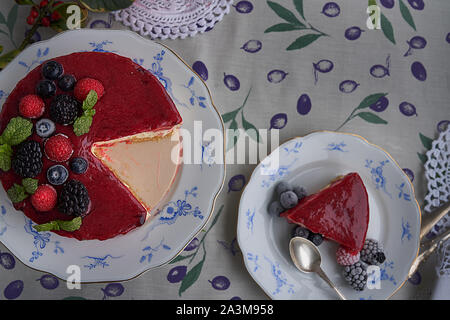 Käsekuchen mit Himbeeren und Brombeeren, Heidelbeeren, Himbeeren und Minze garniert. Auf einem bunten Tischdecke, Blätter und Beeren. Stockfoto