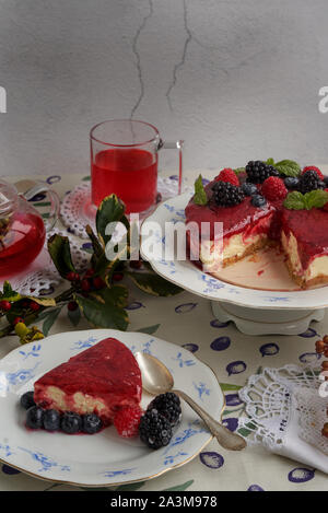 Käsekuchen mit Himbeeren und Brombeeren, Heidelbeeren, Himbeeren und Minze garniert. Auf einem bunten Tischdecke, Blätter und Beeren. Stockfoto