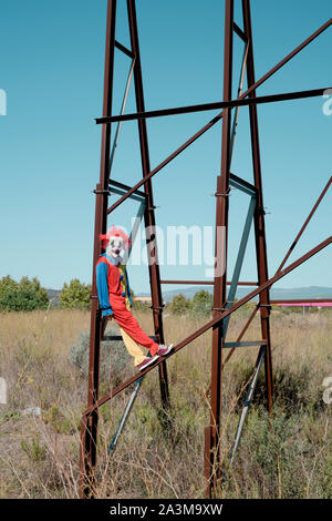 Ein furchtsames Clown trägt einen bunten gelben, roten und blauen Kostüm im Freien, hängen von der rostigen Struktur eines verlassenen Reklametafeln Stockfoto