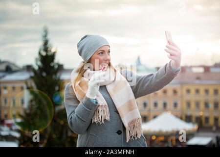 Junge Frau in Weihnachtsmarkt essen Candy Cane und unter selfie. Mädchen auf gestrickte warme Mütze und Schal. Beleuchtete und dekorierte Messe Kiosk, Geschäfte Stockfoto