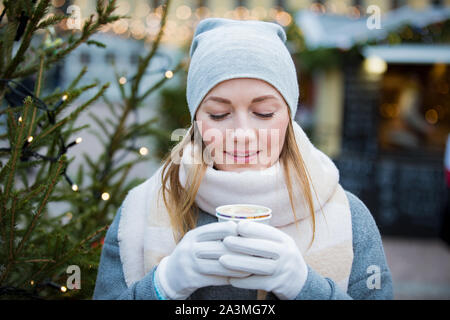 Junge Frau in Weihnachtsmarkt trinken Tasse heiße Schokolade mit Marshmallows tragen gestrickte warme Mütze und Schal. Beleuchtete und Geschäfte eingerichtet Stockfoto