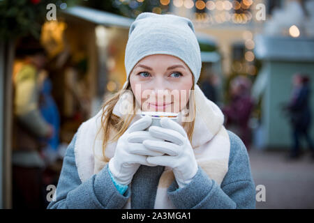 Junge Frau in Weihnachtsmarkt trinken Tasse heiße Schokolade mit Marshmallows tragen gestrickte warme Mütze und Schal. Beleuchtete und Geschäfte eingerichtet Stockfoto