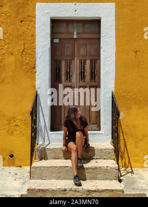 Frau sitzt in einem typischen kolonialen Fassade in Gelb mit Holztür, Izamal (Mexiko) Stockfoto