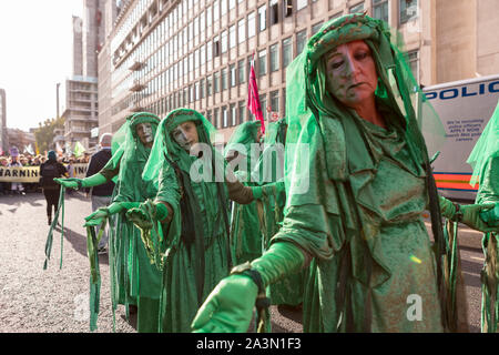 Westminster, London, Großbritannien. Oktober 2019. Mitglieder der Extinction Rebellion-Formation „Green Spirits“ nehmen an einem theatralischen Protest während der Oktober-Rebellion in London teil. Die Darsteller tragen aufwendige grüne Kostüme mit Schleiern und Gesichtsfarbe und Strecken ihre Arme in einer symbolischen Geste entlang der Victoria Street. Hinter ihnen halten andere Aktivisten Banner, während im Hintergrund ein Polizeiwagen zu sehen ist. Die Demonstration verdeutlicht die Dringlichkeit der Klimakrise durch kreative, gewaltfreie Performancekunst in einem geschäftigen städtischen Umfeld. Penelope Barritt/Alamy Live News Stockfoto