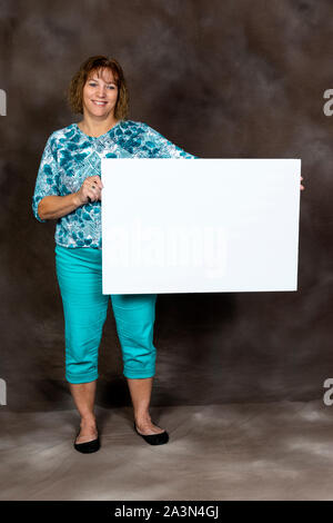 Vertikaler eine freundliche Frau mittleren Alters, die eine leere Zeichen auf braunem Grund. Stockfoto