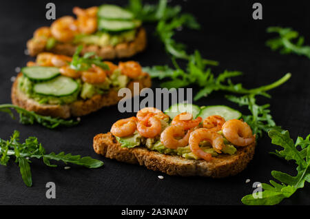 Leckeren snack Toast mit Avocado, gebratene Garnelen, Gurke und Rucola auf schwarzem Hintergrund. Selektiver Fokus, Nahaufnahme Stockfoto