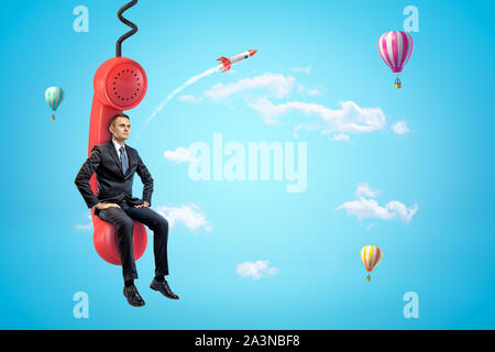 Junge Unternehmer sitzen auf roten Telefonhörer in blauer Himmel mit Heißluftballons und Rakete im Hintergrund. Stockfoto