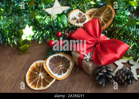 Weihnachten Hintergrund mit dekorativen Zusammensetzung der grünen Lametta, getrocknete Orangen, roten Berres, Pine Cone, Geschenk mit roter Schleife und Sterne auf Holzbrett Stockfoto