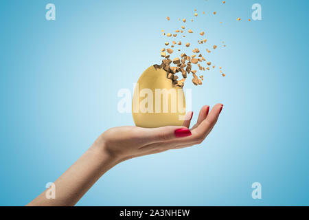 Weibliche Hand, die eine goldene Ei zerbricht in Stücke auf blauem Hintergrund Stockfoto