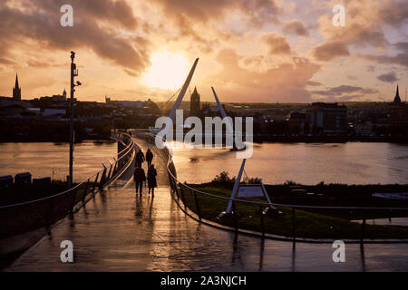 Derry/Londonderry, Nordirland, Großbritannien - 9. Oktober 2019. UK Wetter: Sonne über der Peace Bridge als Fußgänger der Foyle Fluss überqueren. Die Sonne machte eine späte Auftreten nach einer unruhigen Nachmittag. Stockfoto