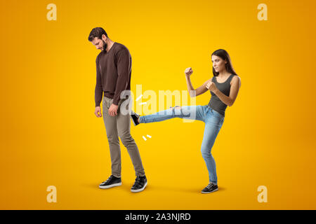 Junge brünette Mädchen mit lässigen Jeans und T-Shirt kicking traurige junge Mann in Freizeitkleidung auf gelbem Hintergrund Stockfoto