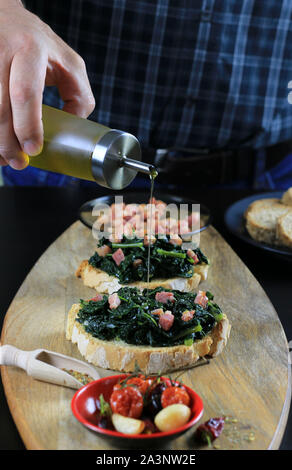 Konzept der italienisches Essen. Nahaufnahme der männlichen Händen gießen Olivenöl über Bruschetta mit schwarzen Kraut und Speck. Rustikale italienische Beilage Stockfoto