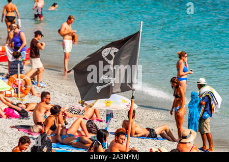 Monterosso al Mare, Italien - September 02, 2019: Schwarz Jolly Roger piraten Flagge am Strand voller sanbathing und Schwimmen Touristen in berühmten Monteros Stockfoto