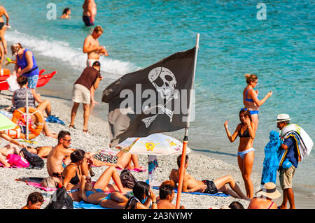 Monterosso al Mare, Italien - September 02, 2019: Schwarz Jolly Roger piraten Flagge am Strand voller sanbathing und Schwimmen Touristen in berühmten Monteros Stockfoto