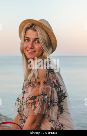 Junge hübsche Frau mit blonden Haaren in rosa Hemd und Hut sitzen auf die Steine in der Nähe vom Meer bei Sonnenaufgang Stockfoto