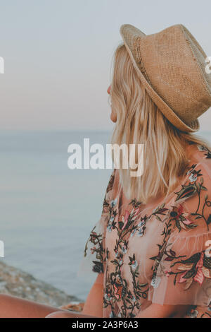 Junge hübsche Frau mit blonden Haaren in rosa Hemd und Hut sitzen auf die Steine in der Nähe vom Meer bei Sonnenaufgang Stockfoto