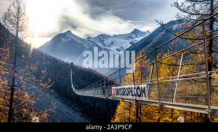 Randa, Schweiz, 8. Oktober 2019: lange Hängebrücke in Zermatt über Landschaft verschneite Berge Wald bei Sonnenuntergang. Suspension Bridge. Lange Brücken. Stockfoto