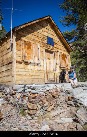 Frau mit Hund sitzt und ruht außerhalb einer Wüste Förster Holzhütte in der Satteltasche See in der östlichen Sierra Nevada Bergen in Kalifornien, USA Stockfoto