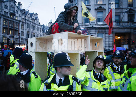 Ein Mitglied des Klimawandels Aktivist Gruppe Aussterben Rebellion (XR) sitzt oben auf einer Holzkonstruktion in Trafalgar Square, ihr Handgelenk Kabel - damit verbunden, von Polizisten umgeben, am Eröffnungstag der lange geplanten "Internationalen Rebellion' in London. Von frühen Dienstag Abend der Metropolitan Police Berichterstattung waren insgesamt 531 Festnahmen so weit über den zwei Tagen der Proteste in der Stadt gemacht worden war, mit Polizisten, damit sich alle Personen und Zelte, die von vielen Seiten von Aktivisten gestern genommen. Ähnliche Blockaden vom Aussterben Rebellion im April, an Standorten inclu Stockfoto