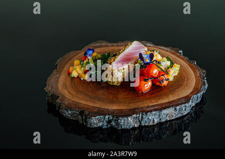 Gebratener Thunfisch Steaks verkrustet mit Kräutern und gegrilltem Gemüse und Mango Salsa auf Holzplatte auf schwarzem Hintergrund Stockfoto