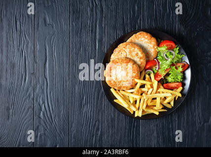 Leckere hausgemachte Türkei Burger serviert mit Salat Tomate Salat und Kartoffeln Pommes frites auf einer schwarzen Platte auf einem schwarzen Holz- Tabelle, Ansicht von oben, flatlay Stockfoto