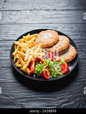 Gebratene Türkei Burger mit Salat Tomate Salat und Pommes frites auf einer schwarzen Platte auf schwarzem Holz Tisch serviert, vertikale Ansicht von oben Stockfoto