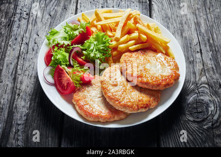 Nahaufnahme der Fried Turkey Burger serviert mit Salat Tomate Salat und Pommes frites auf einem weißen Teller auf einem rustikalen Holztisch, horizontale Ansicht von Abo Stockfoto