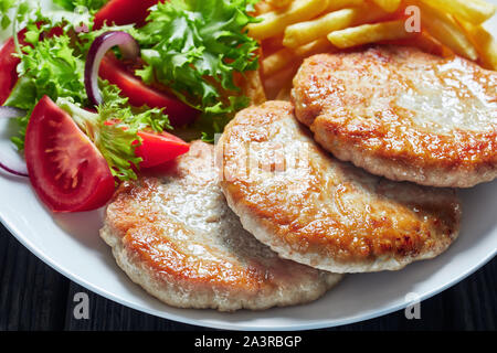 Nahaufnahme der Fried Turkey Burger serviert mit Salat Tomate Salat und Pommes frites auf einem weißen Teller auf einem rustikalen Holztisch, horizontale Ansicht von Abo Stockfoto