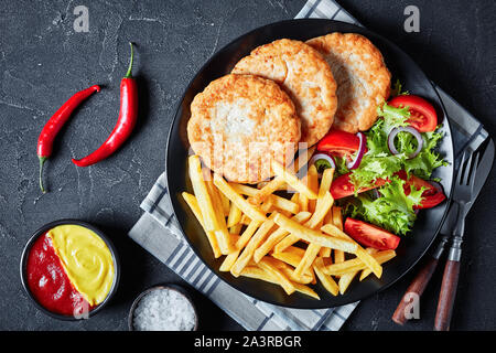 Nahaufnahme der Fried Turkey Burger serviert mit Salat Tomate Salat und Pommes frites auf einer schwarzen Platte auf einer konkreten Tabelle, mit Ketchup und Senf hor Stockfoto