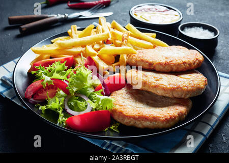 Nahaufnahme der Fried Turkey Burger serviert mit Salat Tomate Salat und Pommes frites auf einer schwarzen Platte auf einer konkreten Tabelle, mit Ketchup und Senf, Ho Stockfoto