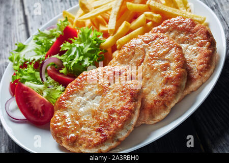 In der Nähe von frisch gebratenen drei Türkei Burger serviert mit Salat Tomate Salat und Pommes frites auf einem weißen Teller auf einem rustikalen Holztisch, horizontal Stockfoto