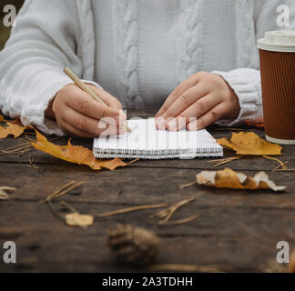 Menschliche Hände mit einer Tasse Kaffee und Schal an den hölzernen Tisch mit Notizbuch und Stift und Blätter im Herbst. Menschliche Hände mit einer Tasse Kaffee und Schal an Stockfoto