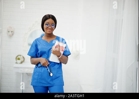 Portrait von Happy weiblichen afrikanischen amerikanischen Jungen Arzt Kinderarzt in blauen Uniform Mantel und Stethoskop mit Büchern in Händen. Gesundheitswesen, Medizin, med. Stockfoto