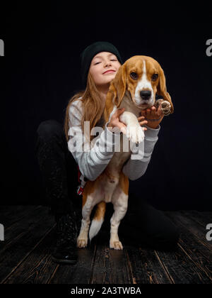 Ein rothaariges Mädchen von zehn Jahre alten spielt mit einem beagle Welpe, sitzend auf einem dunklen Holzboden gegenüber einem schwarzen Hintergrund. Stockfoto