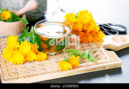 Eine Tasse calendula Infusion Tee mit frischen Ringelblume Blumen und antiken Schere in der Nähe, ein Mörser, pounder mit Kraut und Bettwäsche Threads auf dem backgrou Stockfoto