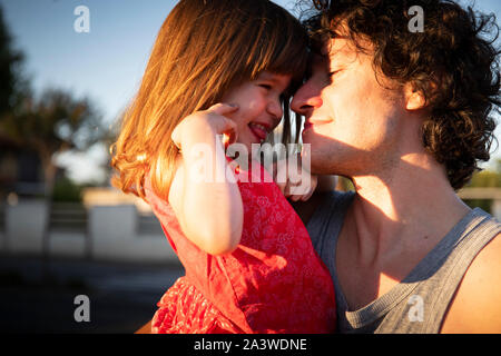 Junger Vater Lachen mit seiner kleinen Tochter bei Sonnenuntergang im Freien. Der Mann schaut sich das Kind und sie trägt ein rotes Kleid. Kopieren Sie Platz auf der Le Stockfoto