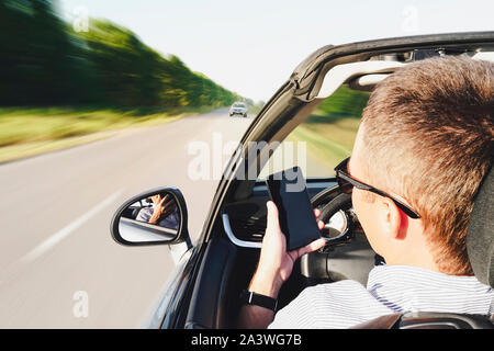 Nahaufnahme von einem Mann mit einem Smartphone während der Fahrt ein Auto. Fahren in den Gegenverkehr. Gefährliche Bewegung. Per Telefon abgelenkt. Der Kerl schreibt ein Chaos Stockfoto